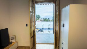 Une pièce avec un plancher en bois, un téléviseur posé sur un meuble, un canapé et une armoire. Une porte donne sur un balcon extérieur doté d’une rambarde et offrant une vue sur des immeubles et des arbres.