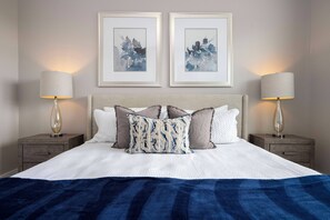 Ein ordentlich bezogenes Bett mit einer weiß-blauen Streifenauflage, zwei gerahmte Blumenbilder an der Wand und zwei Nachttischlampen auf Holznachttischen.