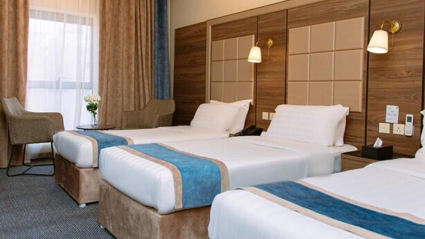A hotel room with two beds, a sitting area with a chair, and a large window with curtains. - GOSONA LBASIKA HOTEL (Madinah)