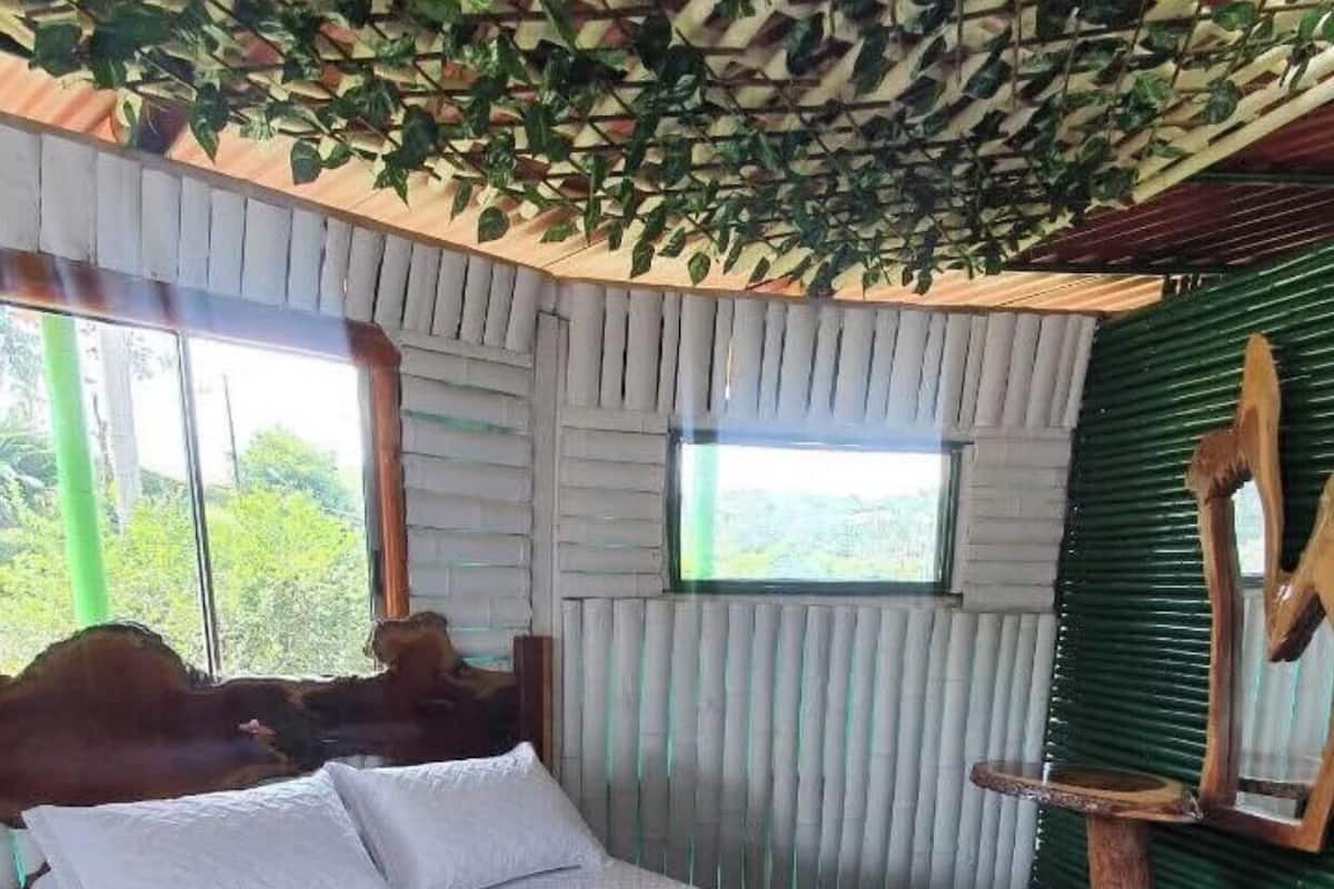 Ein Bett mit weißen Laken und zwei aufgerollten Handtüchern, ein Holz-Kopfteil, ein Fenster mit Blick und eine grüne Wand mit Blätermuster.