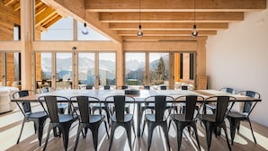 Una gran mesa de madera para comer con sillas, ventanas amplias con vistas a las montañas y un techo alto con vigas a la vista.
