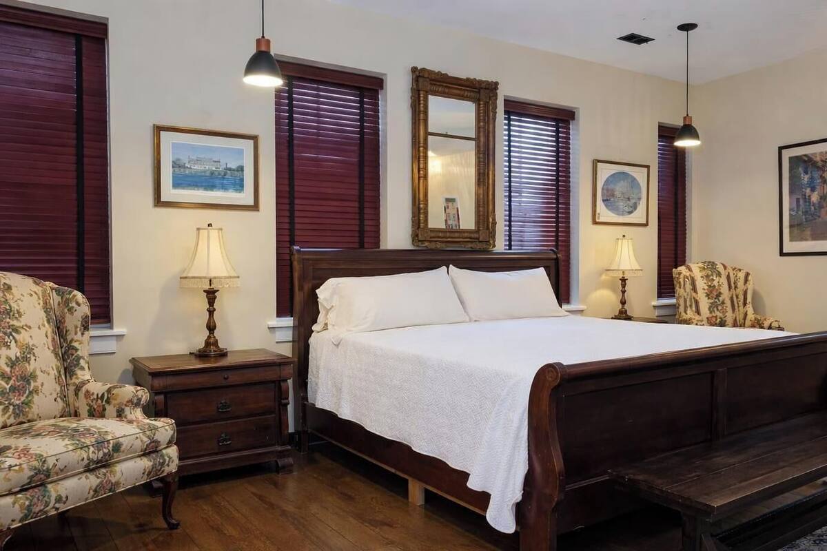 A bedroom with a large bed, wooden headboard, two bedside tables, a chair, and a mirror.
