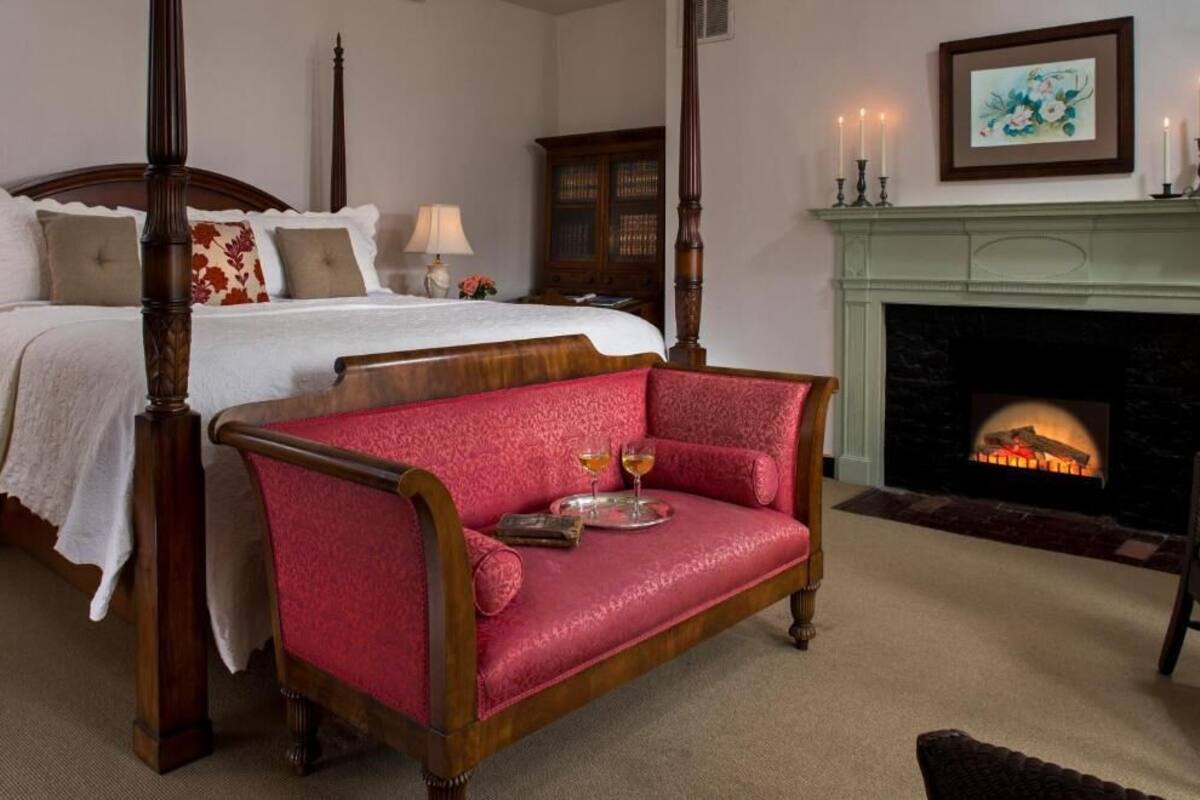 A bedroom with a four-poster bed, a red upholstered sofa, a fireplace, and a framed artwork on the wall.