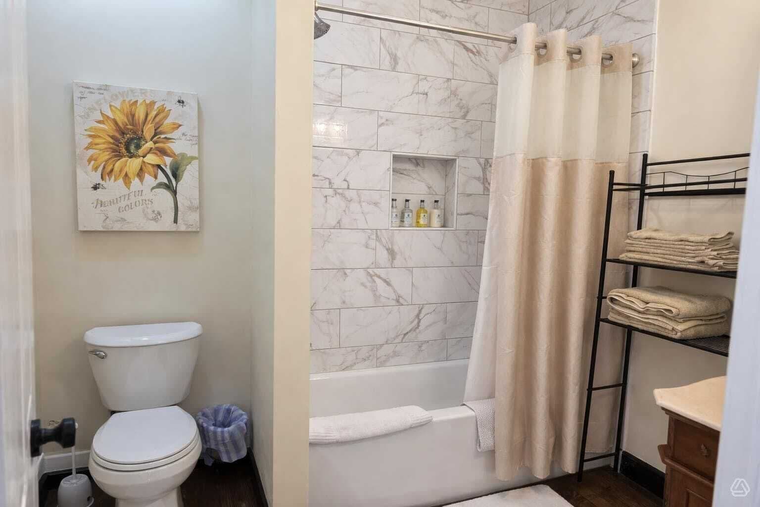 Un baño con inodoro, ducha con pared de mármol, estante con artículos de aseo y toallero.