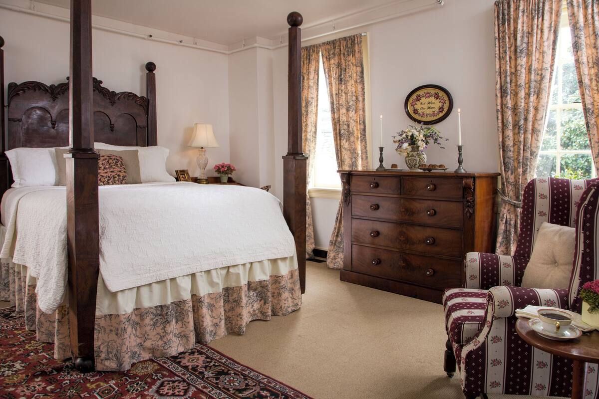 A four-poster bed with a floral bedspread, a wooden dresser, and a patterned armchair in a room with a carpeted floor.
