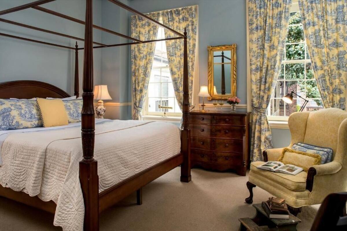 A four-poster bed with a wooden frame, a bedside table with a lamp, a chair with a cushion, and a dresser with a mirror.