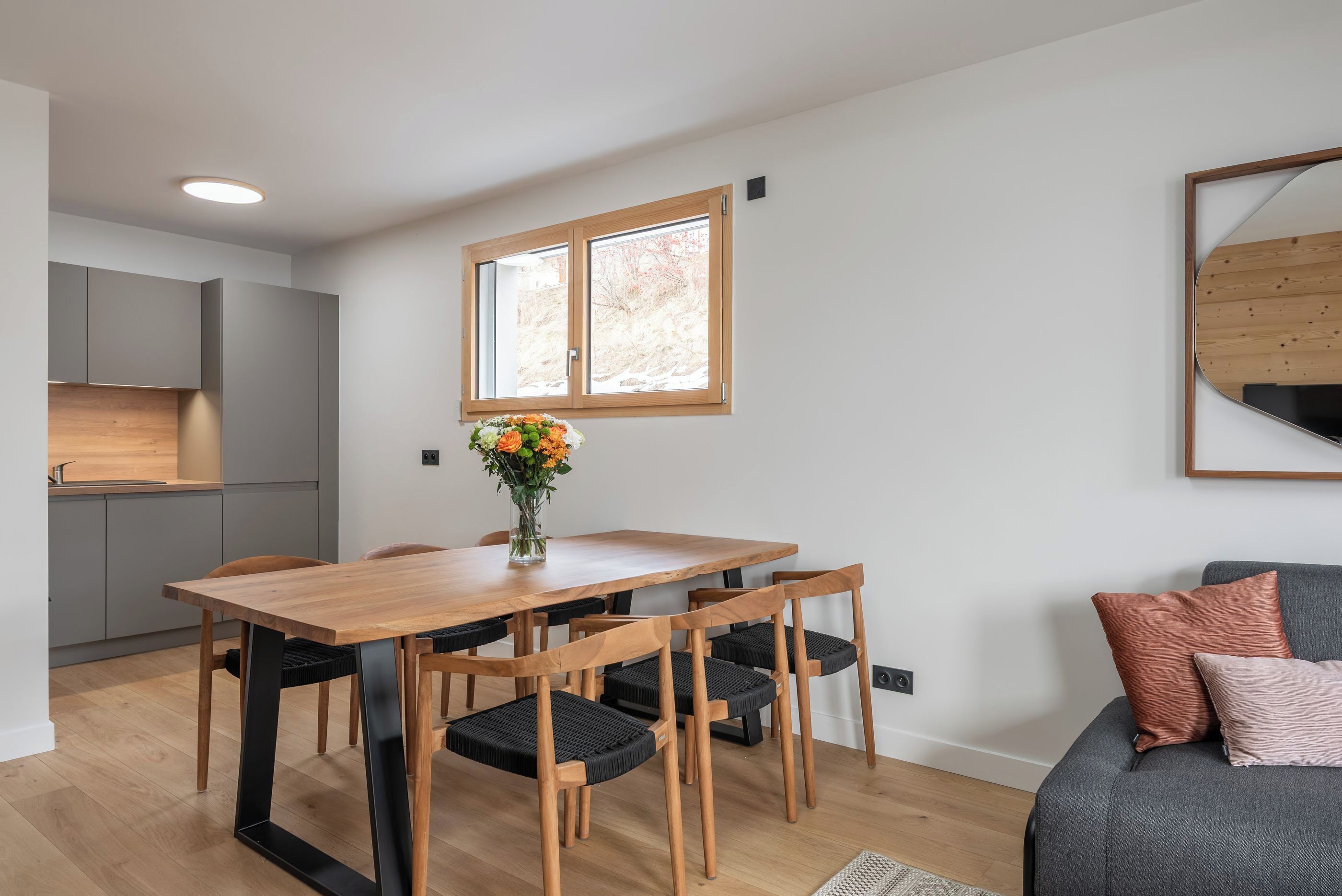 A modern kitchen with grey cabinets, a wooden dining table with four chairs, a window with a view of trees, and a grey sofa with cushions.