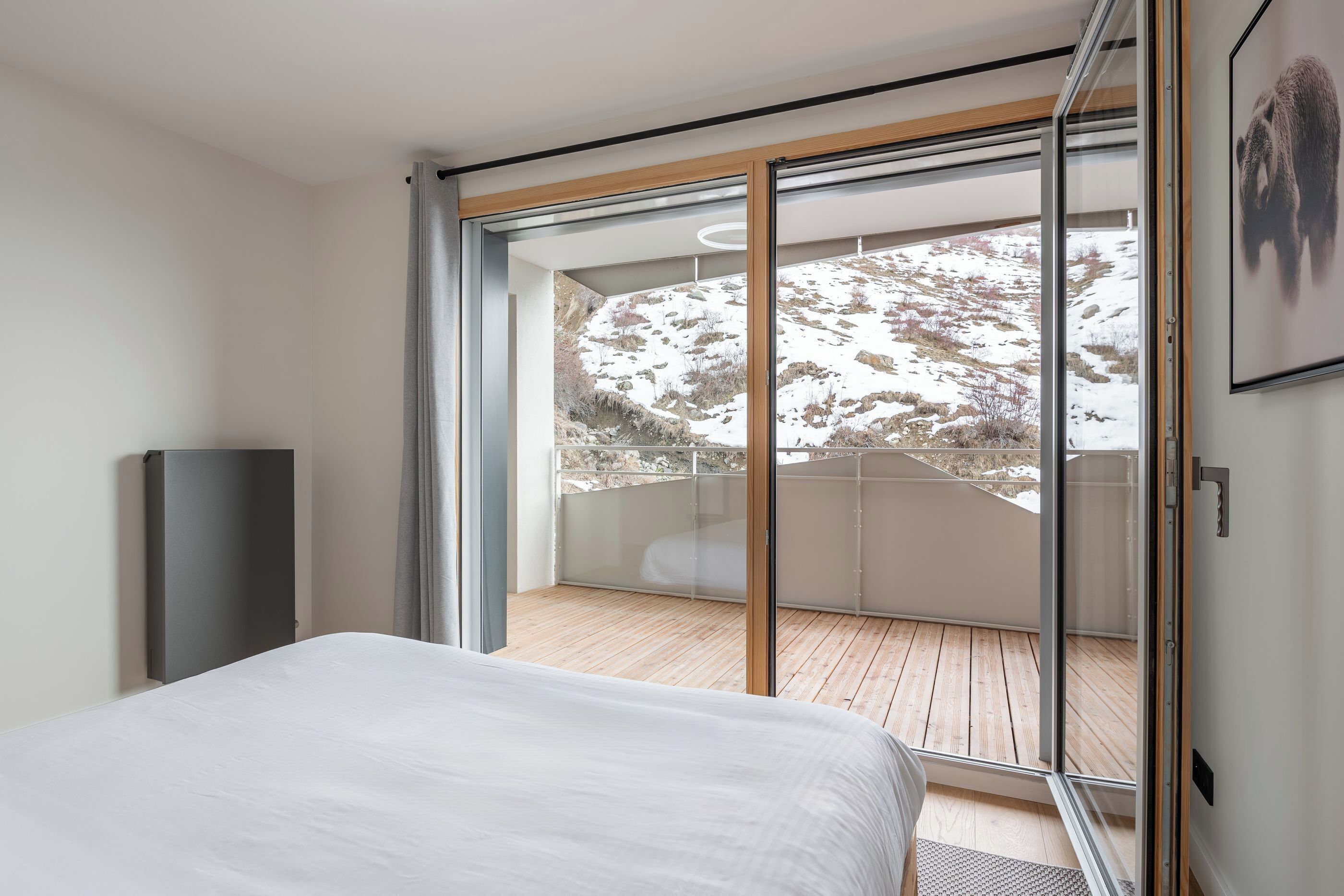 A modern bedroom with a large bed, a sliding glass door leading to a balcony with a view of a snowy landscape.