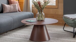Un salon moderne avec un canapé gris, une table basse ronde ornée d’un vase, une lampe, et une vue sur un paysage enneigé à travers de grandes fenêtres.