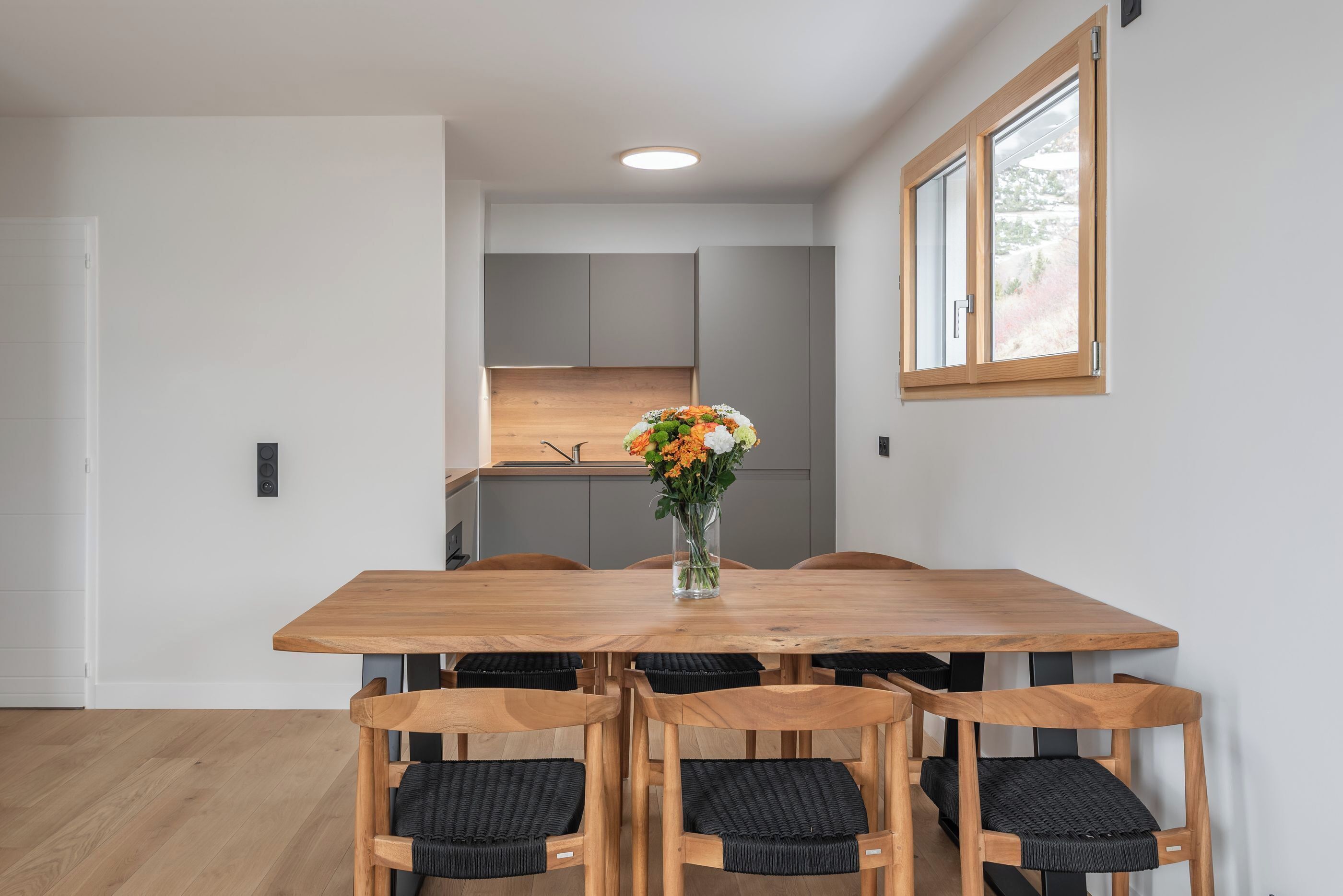 Een eetgedeelte met een houten tafel en stoelen, een vaas met bloemen en een uitzicht op een keuken op de achtergrond.