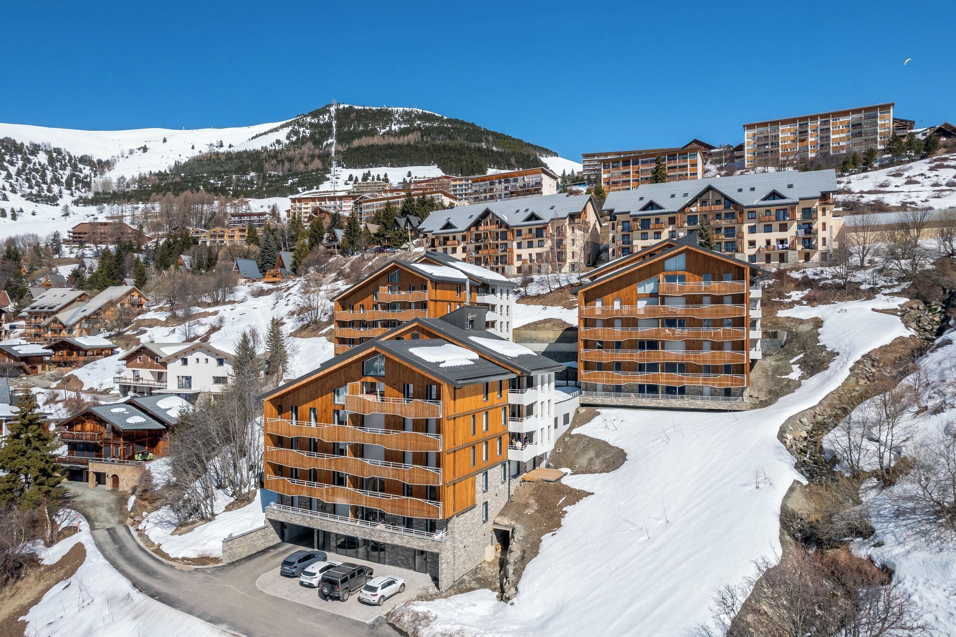 A ski resort with wooden buildings, a parking area, and a snowy mountain backdrop.