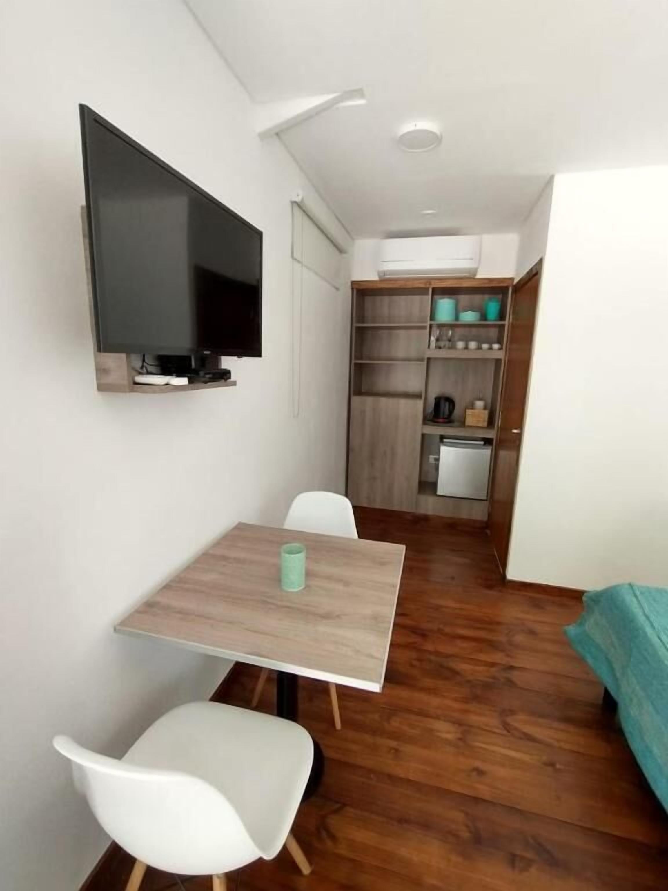 Una habitación compacta con una mesa de comedor de madera, sillas blancas, un televisor de pantalla plana montado en la pared y una pequeña cocina con armarios y microondas.
