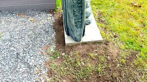 En statue af en skægget figur med hat, der står på en betonfundament, placeret på et træterrasse med græsplæne og et vandareal i baggrunden.