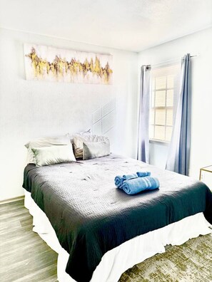 Un dormitorio con cama, almohadas, una colcha, una toalla enrollada, un cuadro en la pared y una ventana con cortinas.