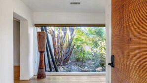 Un interior moderno con suelo de madera, un ventanal con vistas a plantas tropicales y una escultura alta y texturizada.