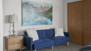 Et værelse med en blå sofa, et trænatbord med en lampe og et stort maleri af et bjerglandskab på væggen.