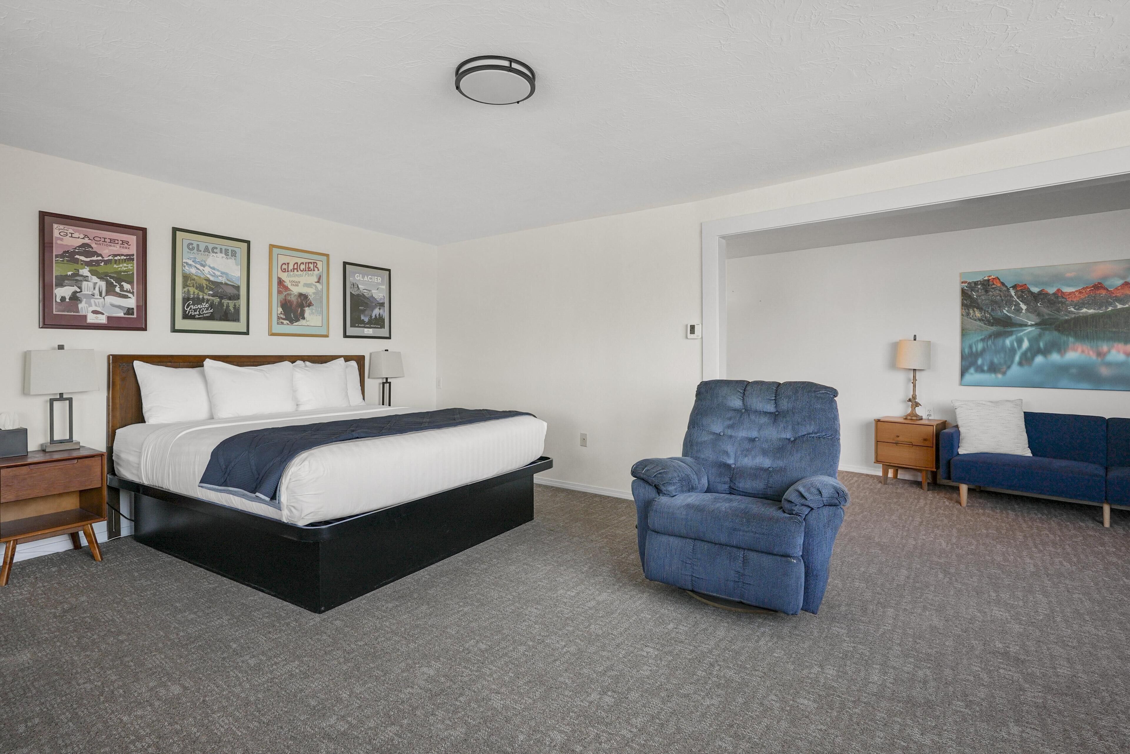 Une chambre d’hôtel avec un lit, un fauteuil bleu, un canapé, une table de chevet, une lampe et quatre tableaux encadrés accrochés au mur.