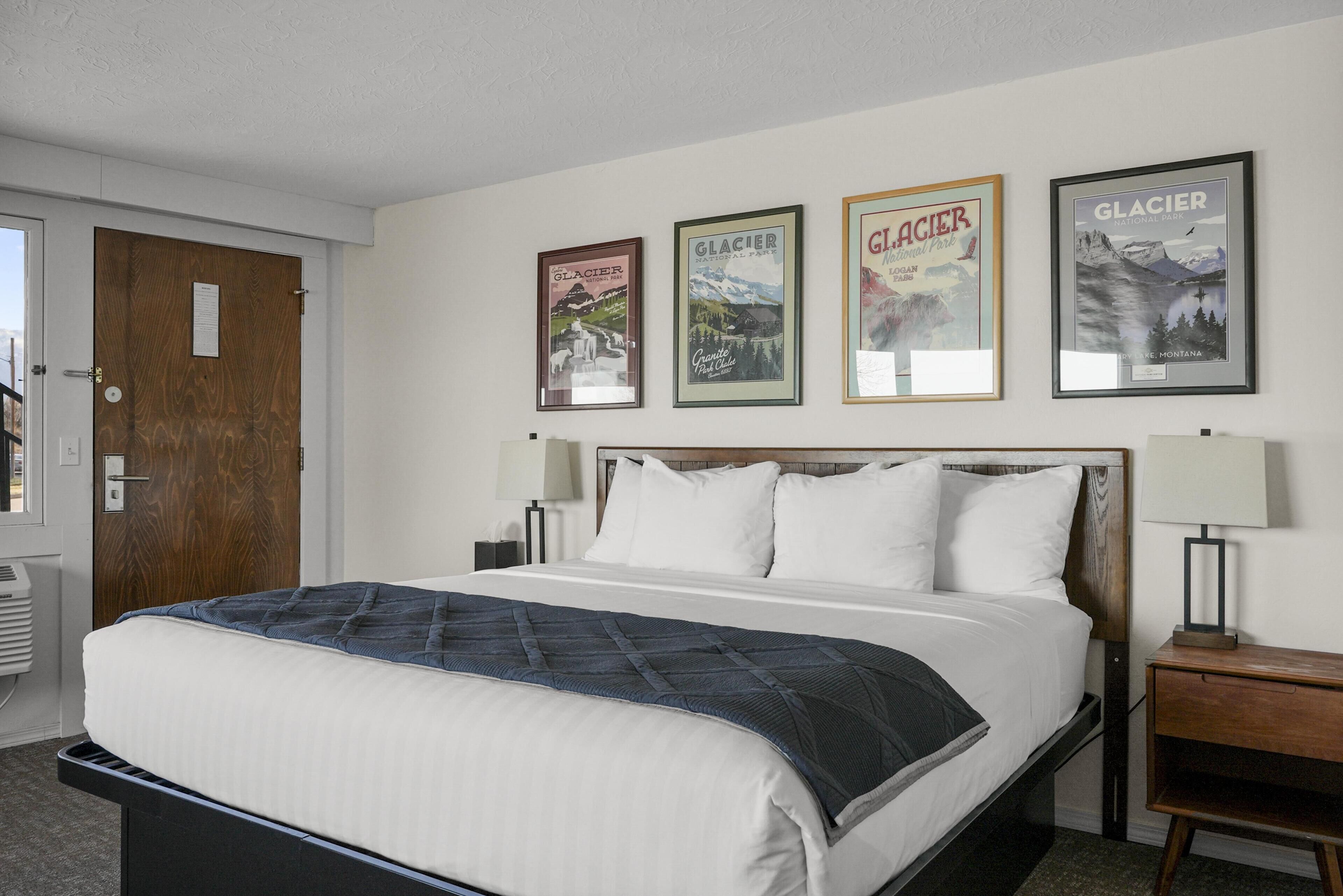 Quarto de hotel com uma cama, uma porta de madeira, uma mesa de cabeceira, um abajur e três quadros na parede.