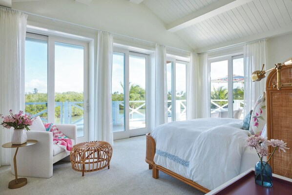A spacious bedroom with a large bed, a sitting area with a chair and a small table, and a view of a balcony with outdoor furniture.
