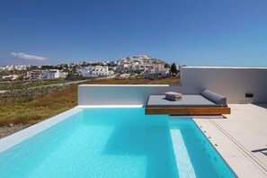 Una piscina a sfioro sul tetto con vista su una cittadina costiera, una panca di legno con cuscini e un cielo azzurro limpido.