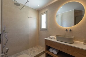 A modern bathroom with a shower, a sink with a basin, a mirror, and a wooden vanity. - Evira Suites (Santorini)