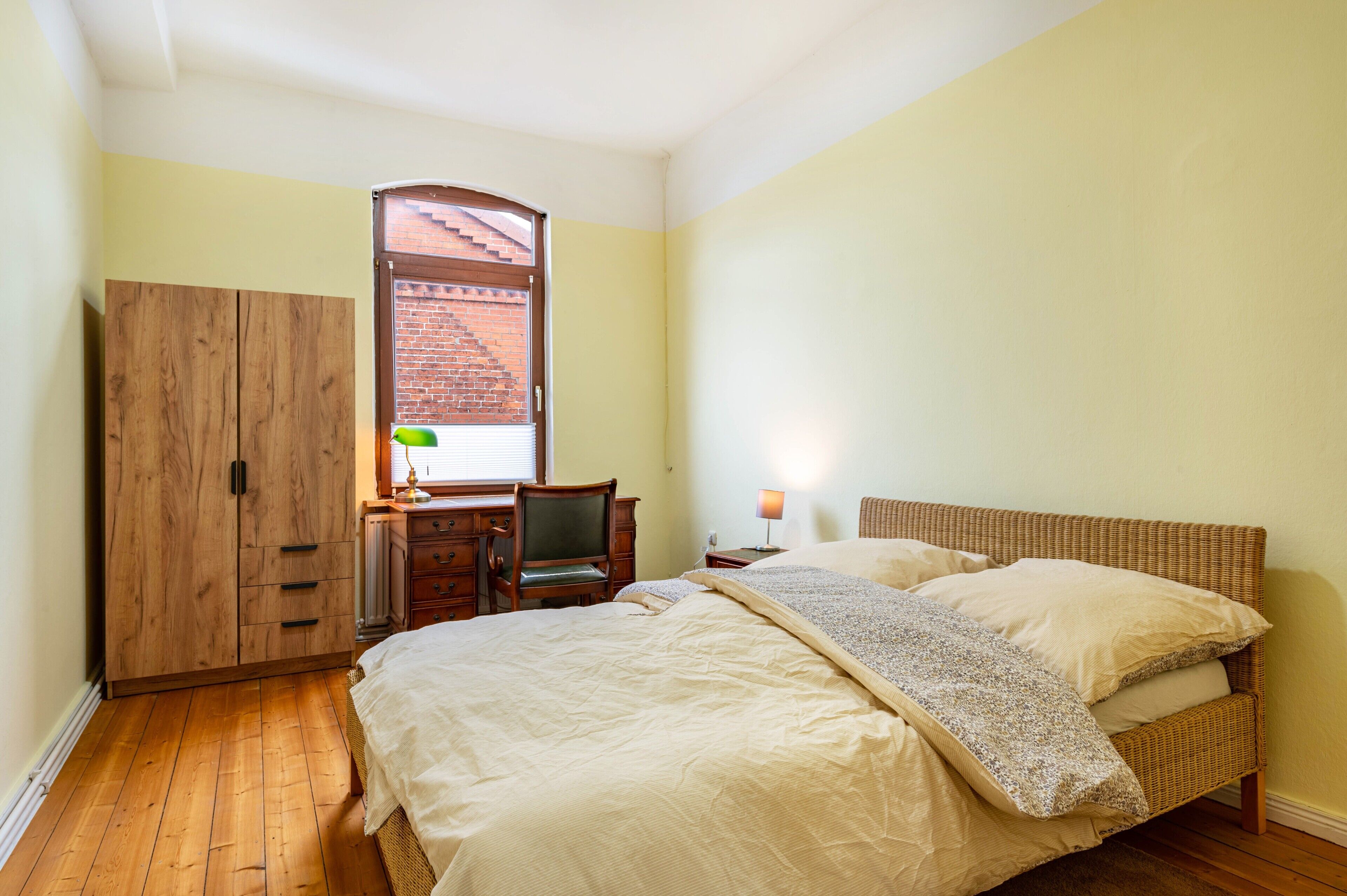 Une chambre à coucher comprenant un lit, une armoire en bois, un bureau, une chaise, une lampe et une fenêtre donnant sur un immeuble en briques.