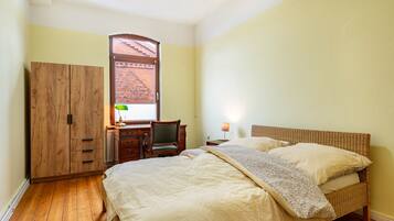 Une chambre à coucher comprenant un lit, une armoire en bois, un bureau, une chaise, une lampe et une fenêtre donnant sur un immeuble en briques.