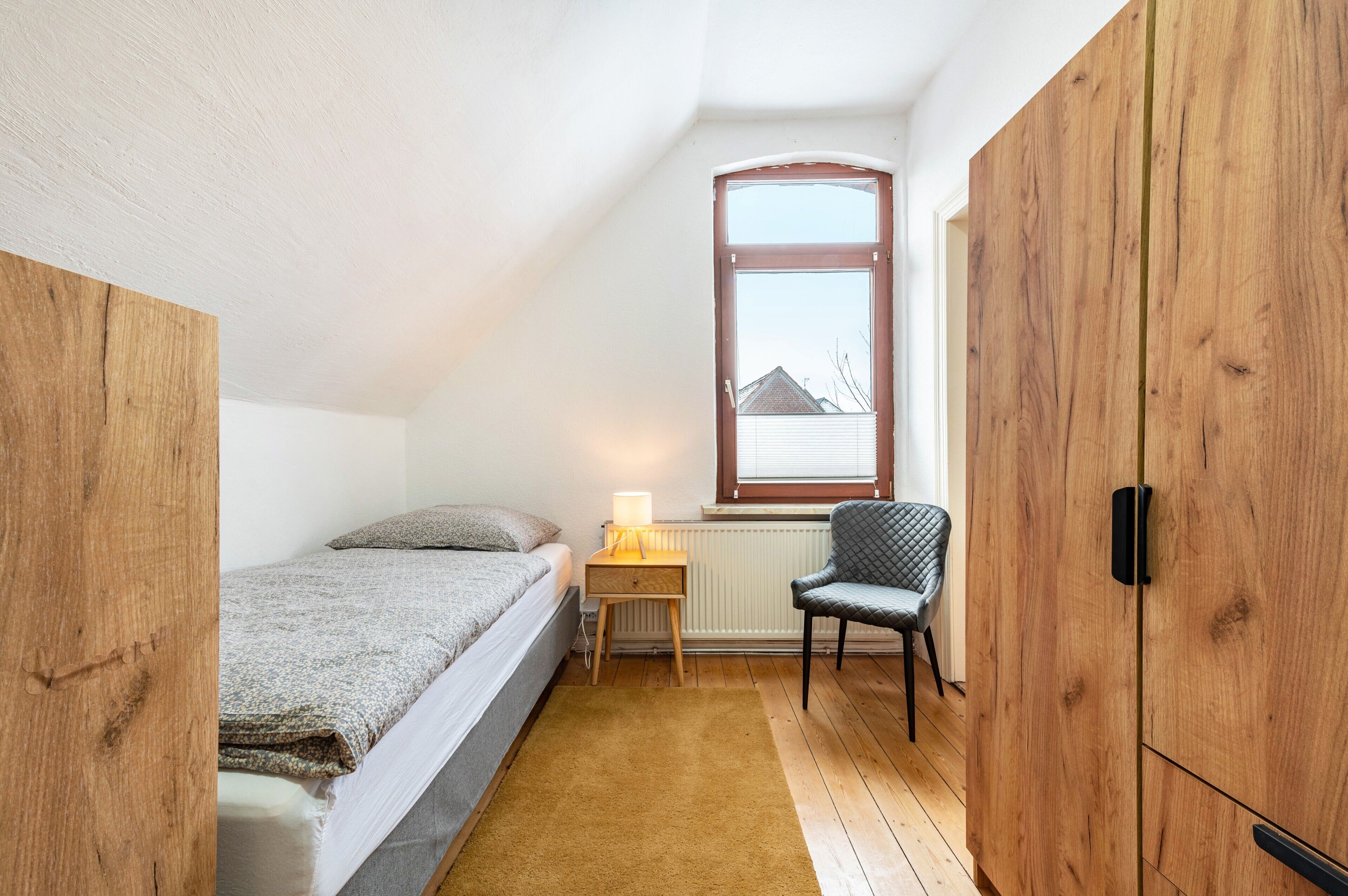 Una camera da letto con un letto, una sedia, una finestra e un armadio in legno.