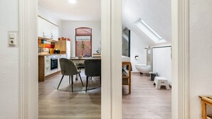 Een moderne keuken met houten kastjes, een eethoek met een ronde tafel en stoelen, een klein krukje, en een dakraam in de aangrenzende badkamer.