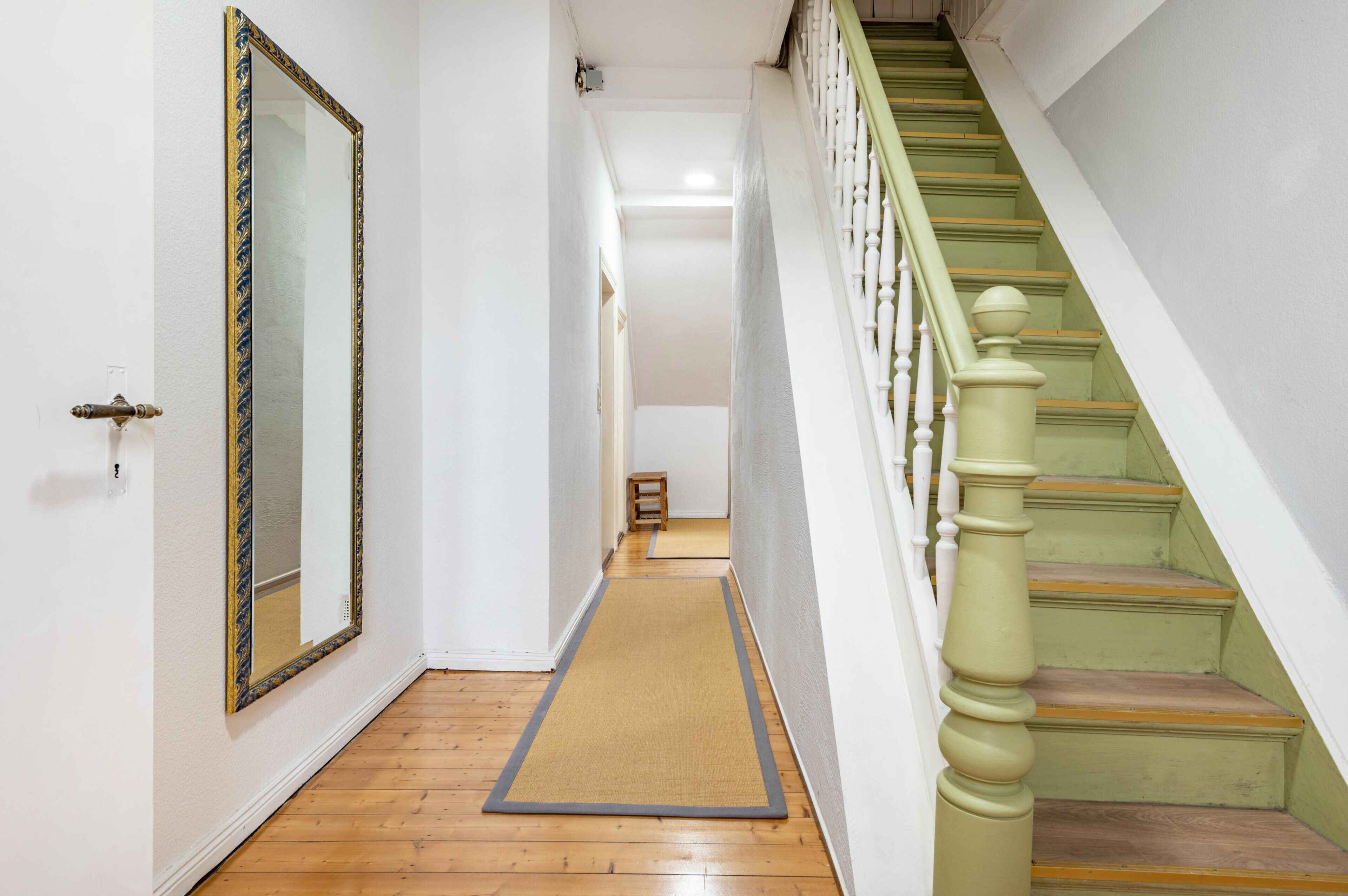 A hallway with a wooden floor, a doormat, a mirror, and a staircase with green steps.