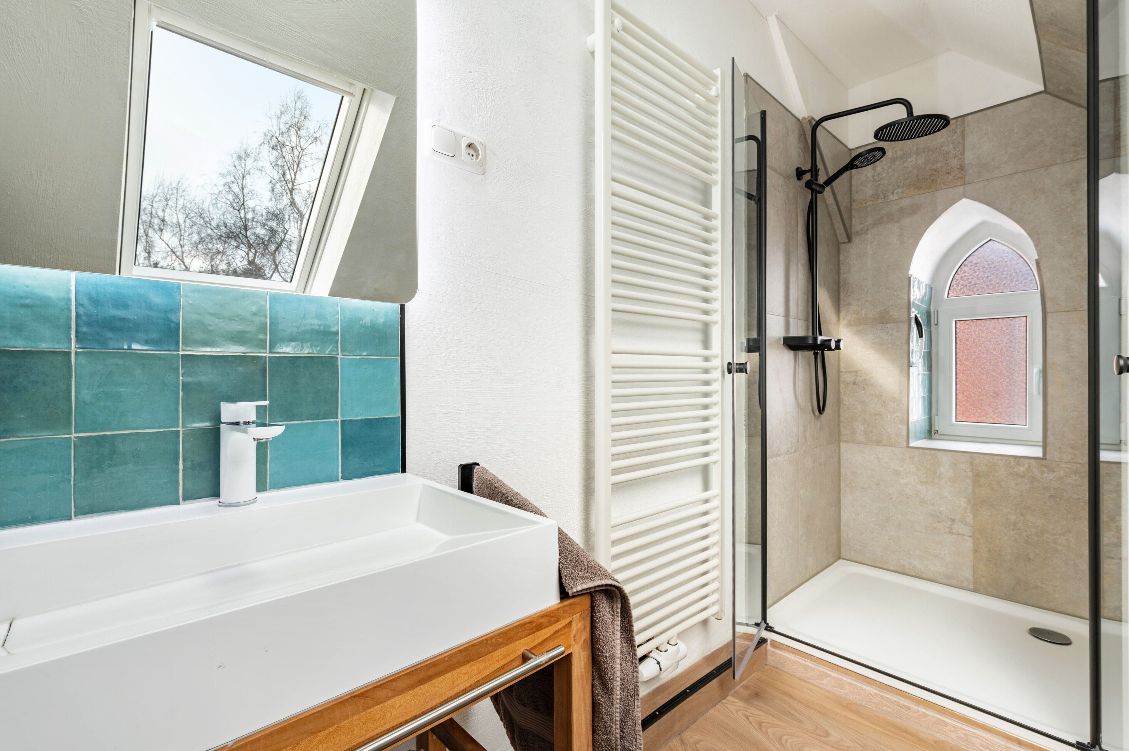 A bathroom with a white sink, a shower with a glass door, and a window with a view of trees.