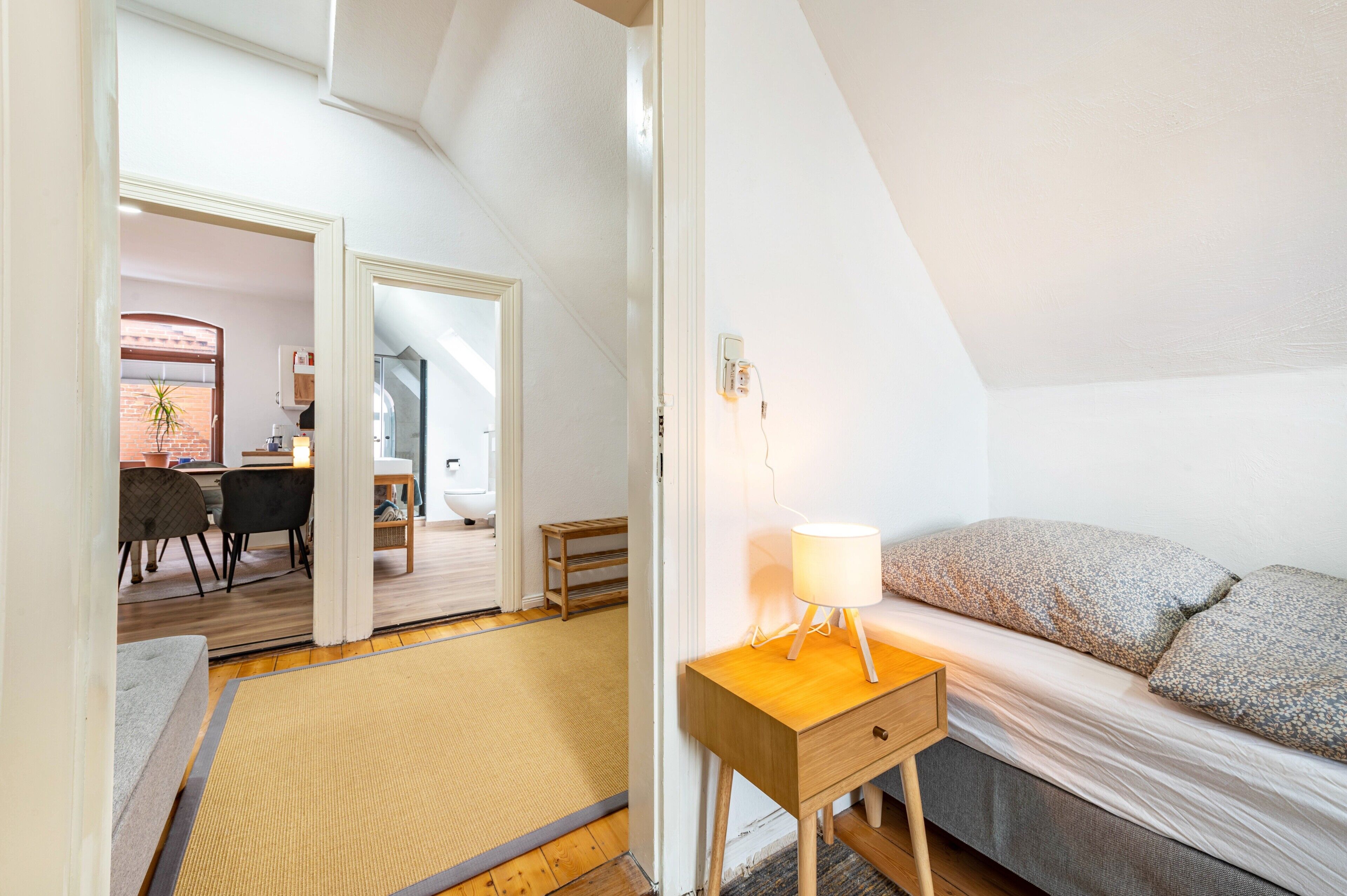 Quarto com cama, mesa de cabeceira, abajur e vista para uma área de jantar com cadeiras e uma mesa.