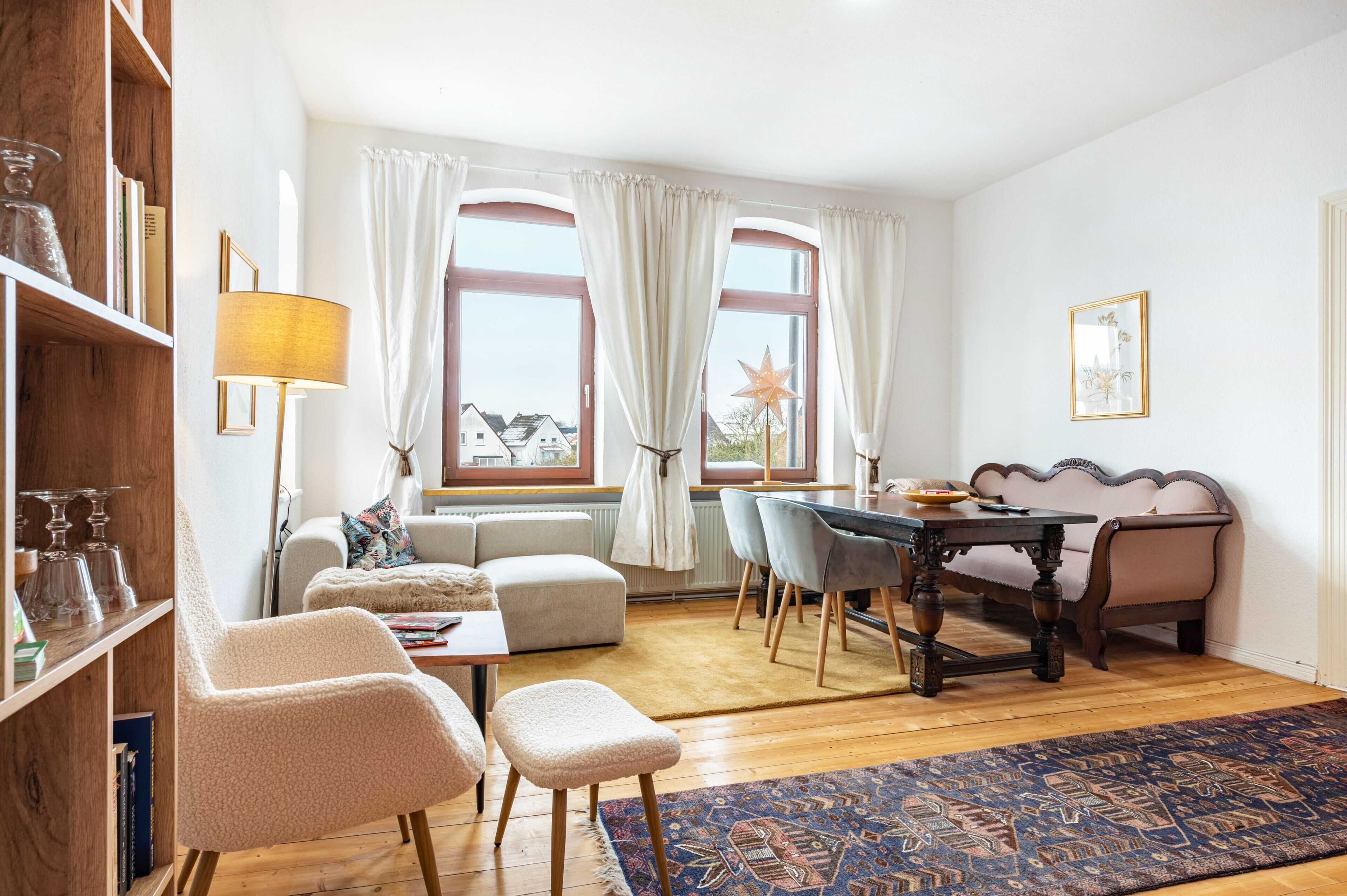 Ein Wohnzimmer mit Sofa, Sessel, Esstisch und Stühlen. Ein Bücherregal, eine Lampe und eine Stern-Dekoration befinden sich am Fensterbrett.