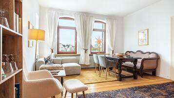 Ein Wohnzimmer mit Sofa, Sessel, Esstisch und Stühlen. Ein Bücherregal, eine Lampe und eine Stern-Dekoration befinden sich am Fensterbrett.