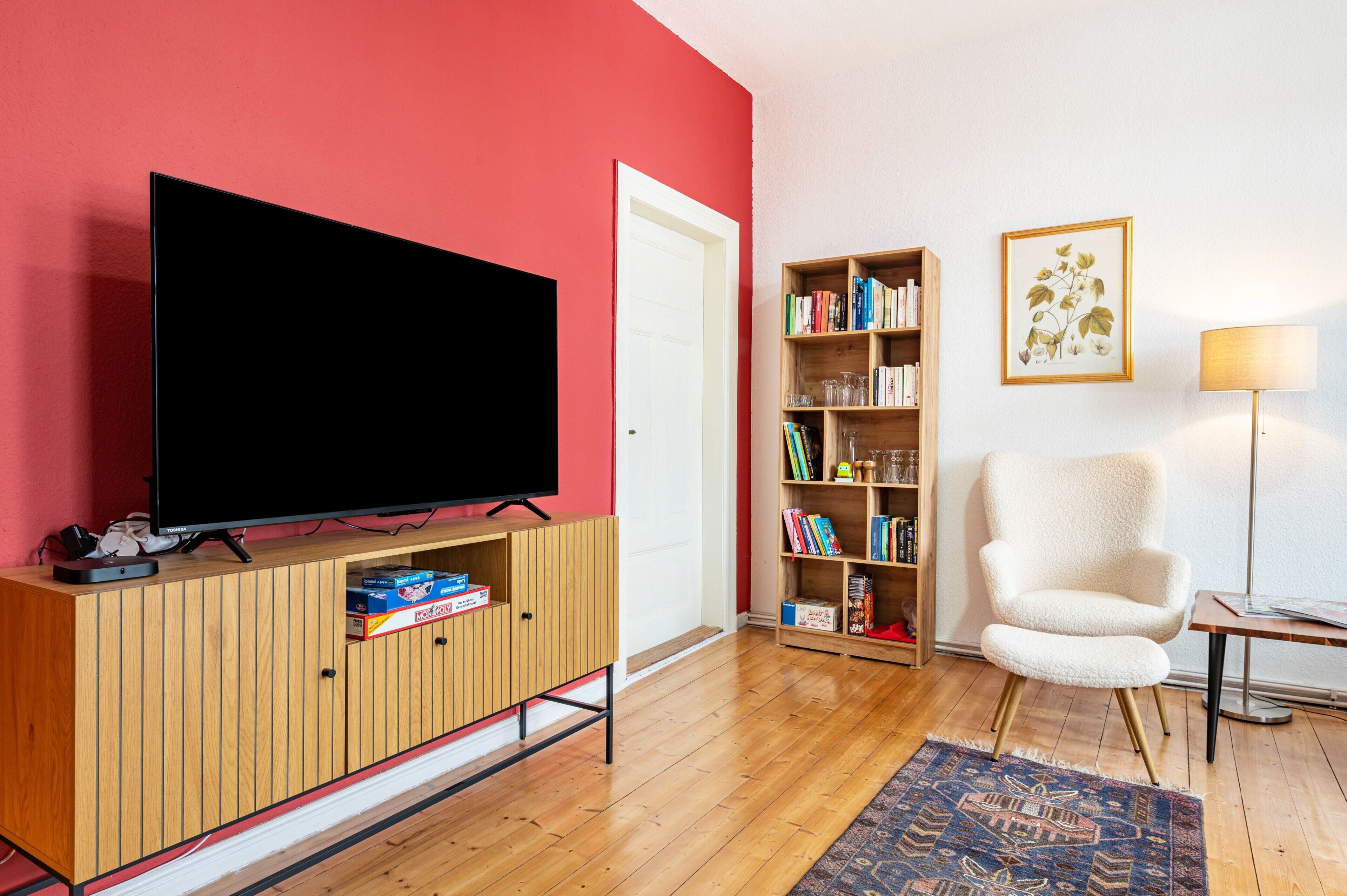 A living room with a flat-screen TV on a wooden cabinet, a bookshelf, a white armchair, and a patterned rug.