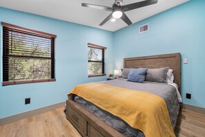 Une chambre à coucher avec un lit, deux fenêtres, un ventilateur de plafond et une table de chevet avec une lampe.