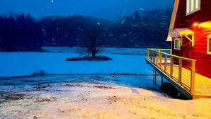 A wooden cabin with a deck, situated by a frozen lake with snow-covered ground and trees in the background.