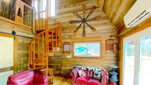 A wooden cabin interior with a red leather chair, a patterned sofa, a wooden staircase, and a ceiling fan.