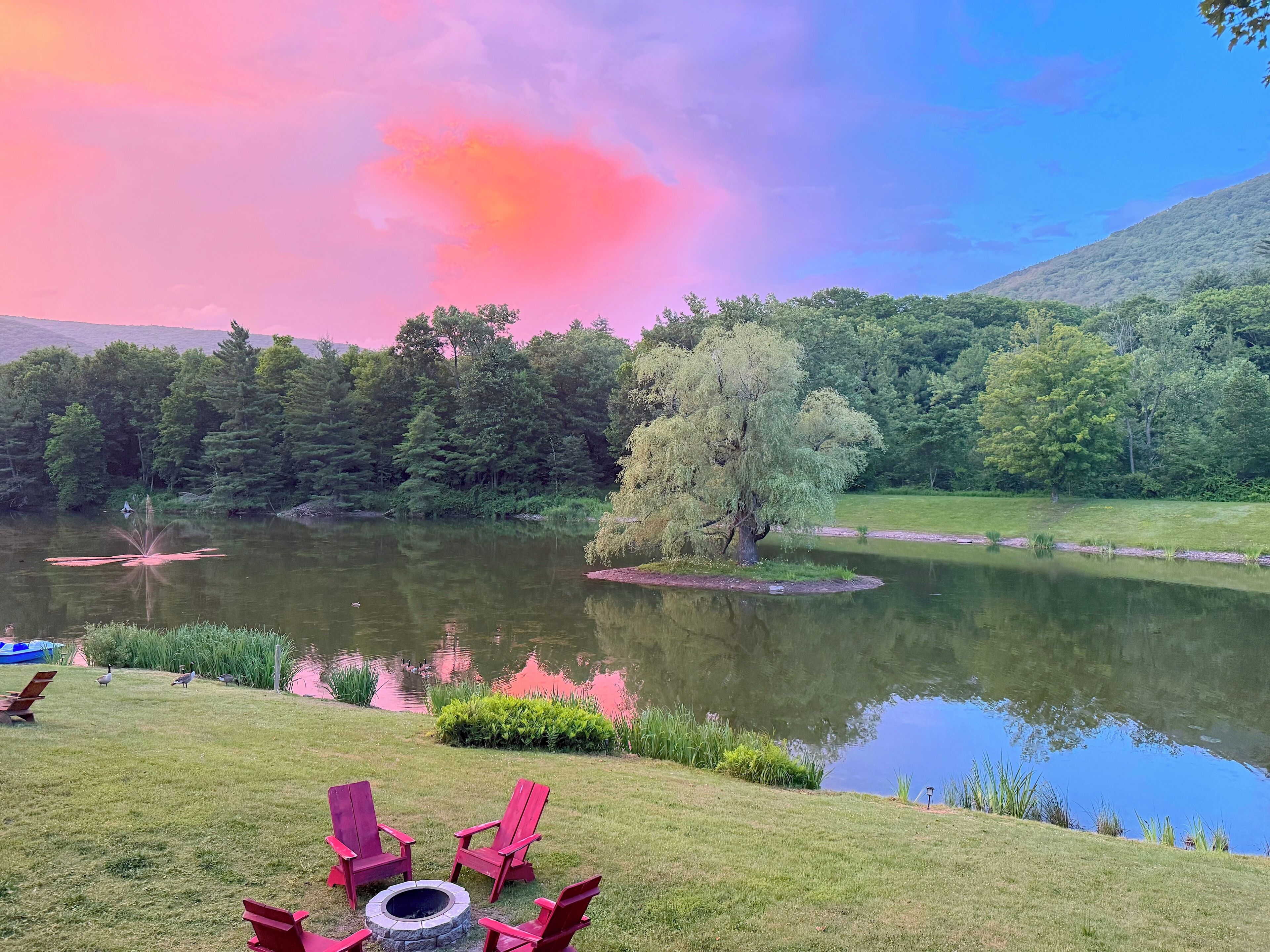 Vista al lago con zona verde, una fogata y sillas rojas.