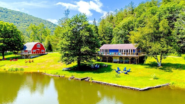 Une propriété au bord du lac, avec une grange rouge et une maison bleue dotée d’une terrasse, le tout entouré d’une végétation luxuriante et sous un ciel dégagé.