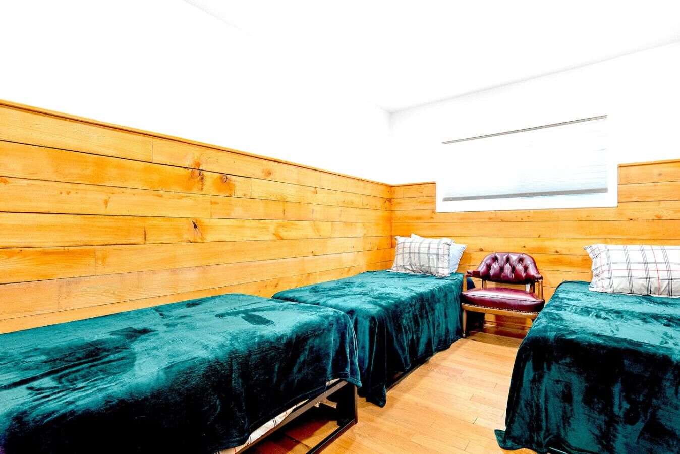 Een kamer met houten wanden en een raam. Er staan twee bedden met groene dekens en een rode stoel.