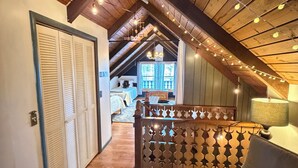 Une chambre aménagée dans les combles, avec des poutres apparentes, des guirlandes lumineuses, un lit et une vue sur le salon.