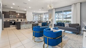 A modern living room with a grey sofa, blue armchairs, and a coffee table. There is a kitchen area with dark cabinets and a stainless steel oven. The room has large windows and a chandelier. - 1627 Exclusive house near Disney 5BD/15P (Davenport)