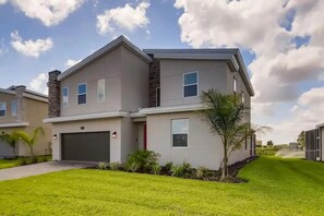 A modern two-story residential building with a garage, a small front yard with grass and plants, and a clear sky. - 9BR Vacation Home Near Disney | Sleeps 24 (Kissimmee)