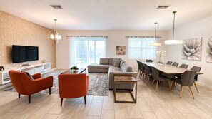 A modern living room with a sofa, chairs, a coffee table, a TV, and a dining area with a table and chairs. - 9BR Vacation Home Near Disney | Sleeps 24 (Kissimmee)