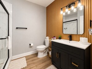 Ein Badezimmer mit Toilette in Weiß, schwarzen Schränken, einer Holzvertäfelung und einem Spiegel mit Beleuchtung.