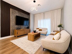 Ein modernes Wohnzimmer mit Flachbildfernseher, Holzmöbeln, einer Couch und einem großen Fenster mit Vorhängen.