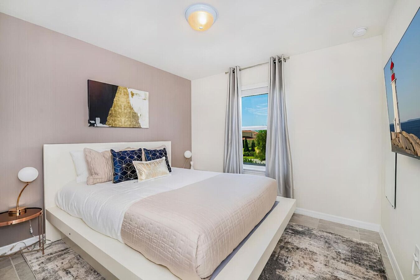 A bedroom with a bed, a nightstand, a lamp, a painting, and a window with curtains.