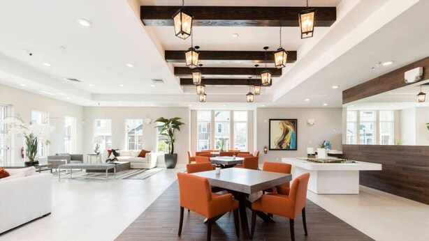 A spacious, modern hotel lobby with a central dining area featuring orange chairs and a large table, surrounded by large windows and a wooden ceiling. - 7532 Perfect Home 5 Minutes from Disney – 5BD 15P (Kissimmee)