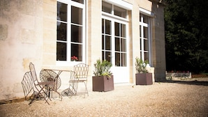 A building with large windows, outdoor seating with metal chairs, and planters with greenery.